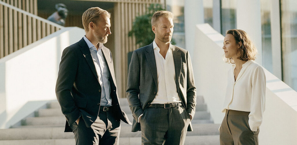 Tre personer i forretningsantrekk står og snakker sammen på en trapp i et lyst og moderne kontorbygg.